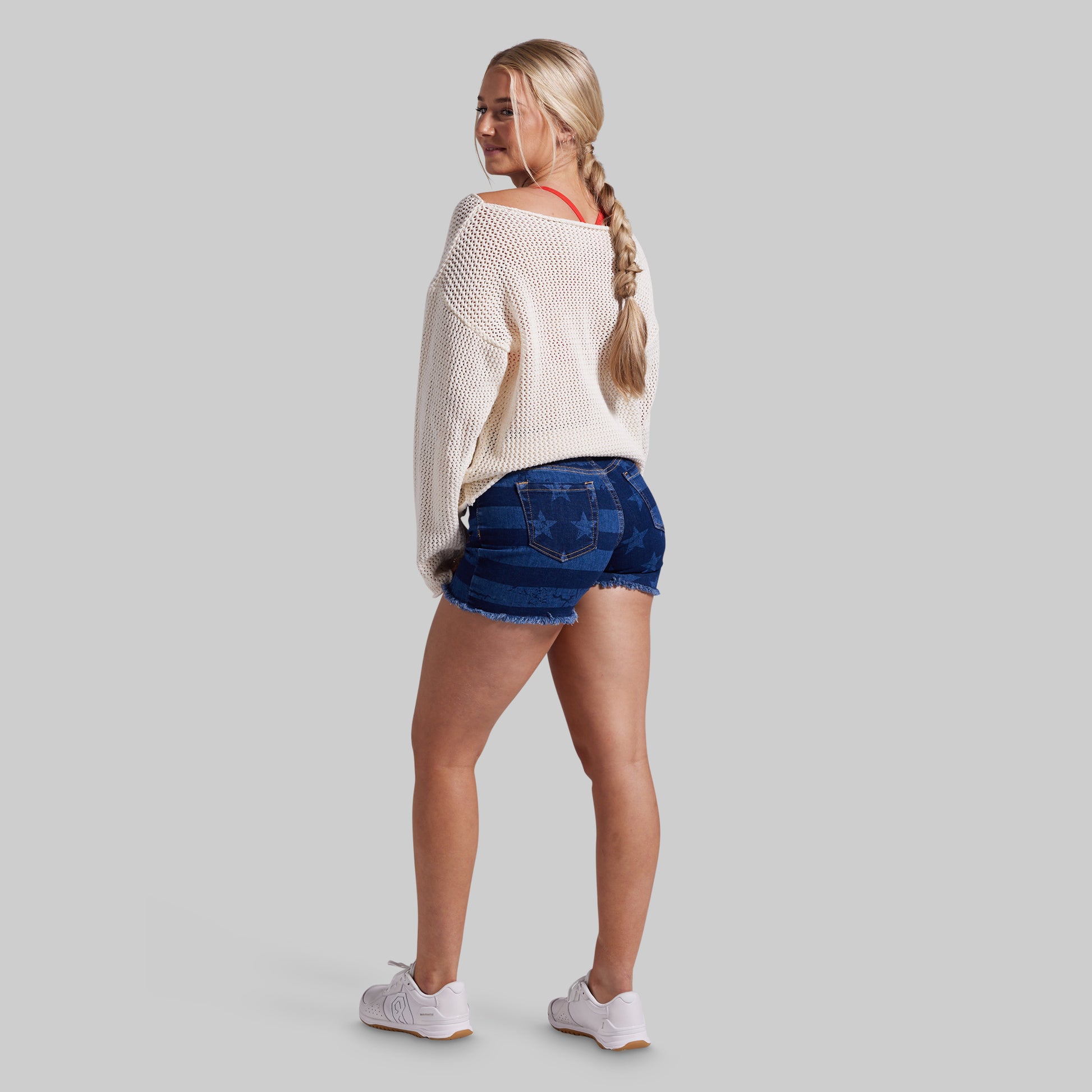 A woman is shown from the back wearing a cream knit sweater, blue denim shorts, and white sneakers against a solid gray background.