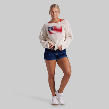A woman modeling a cream knit sweater with an American flag graphic on the chest, paired with blue denim shorts and white sneakers, against a solid gray background.