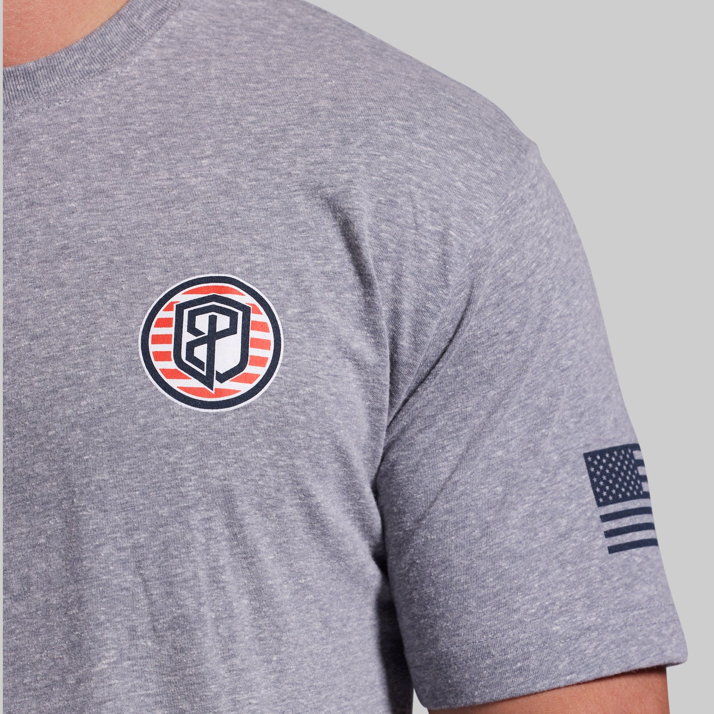 Close-up of a heather gray short-sleeve t-shirt worn by a model, featuring a circular chest logo with red and navy stripes and a navy American flag graphic on the sleeve.