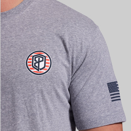 Close-up of a heather gray short-sleeve t-shirt worn by a model, featuring a circular chest logo with red and navy stripes and a navy American flag graphic on the sleeve.