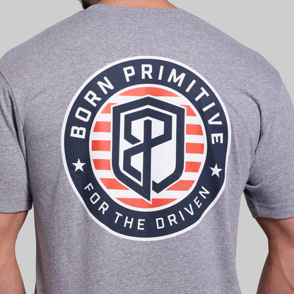 A close-up of a heather gray t-shirt showing the back with a large circular graphic. The graphic features the text 'BORN PRIMITIVE FOR THE DRIVEN' in white around a shield logo with red and white stripes and a navy blue 'BP' monogram in the center.