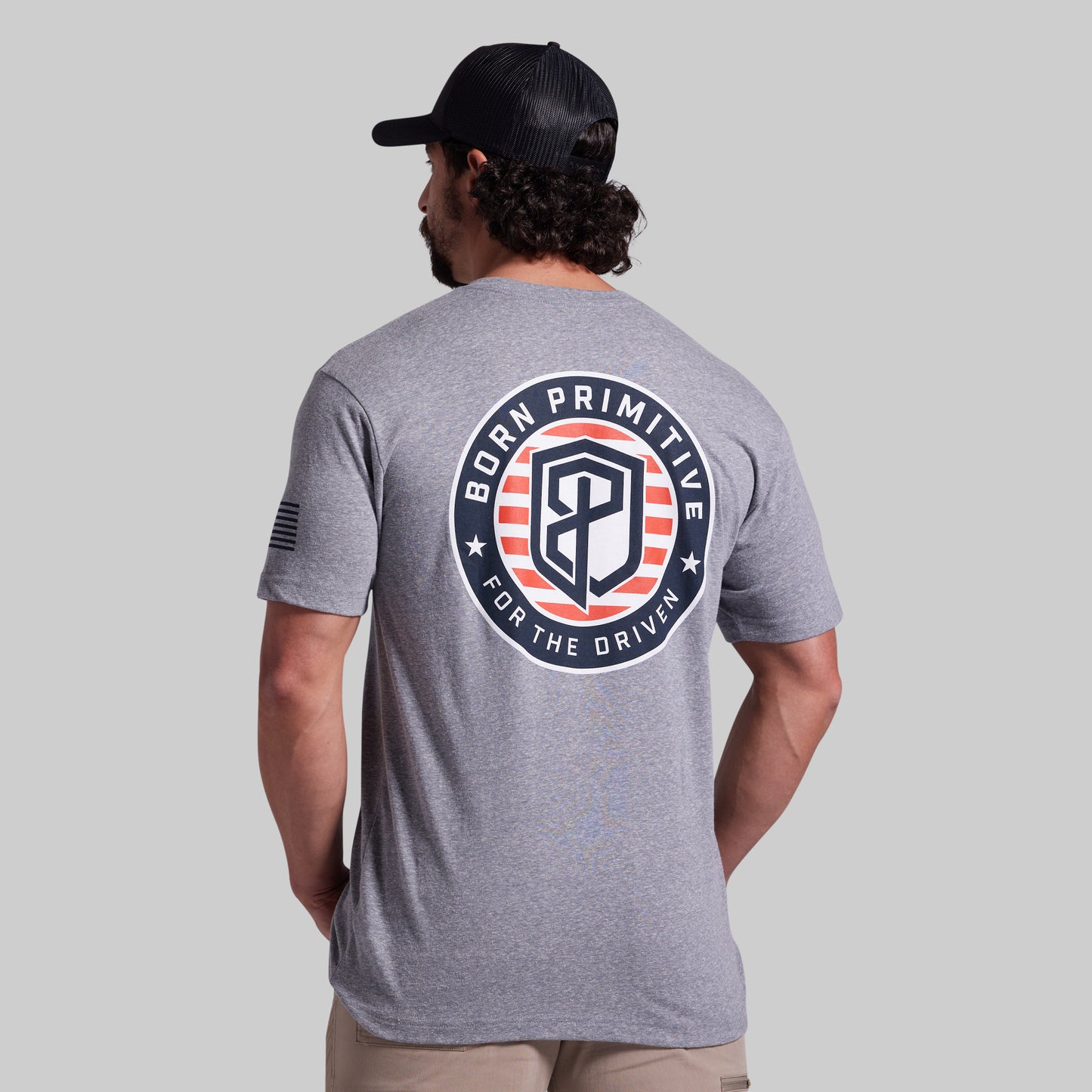 A man wearing a heather gray t-shirt from the back, featuring a large circular 'Born Primitive For The Driven' graphic in navy, white, and red on the back. He is also wearing a black baseball cap and khaki pants, set against a solid light gray background.