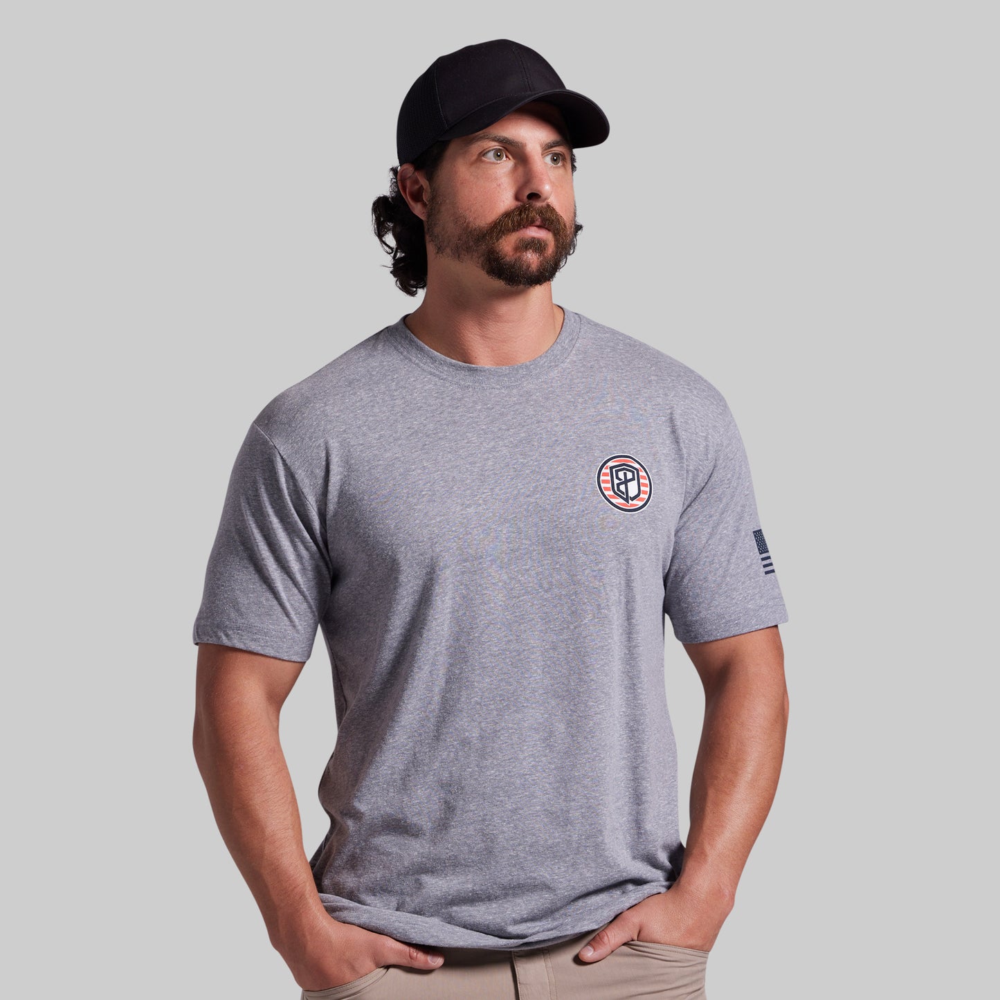 A man wearing a heather gray crewneck t-shirt with a circular logo on the left chest and a small flag print on the sleeve, paired with a black baseball cap and khaki pants, set against a solid gray background.