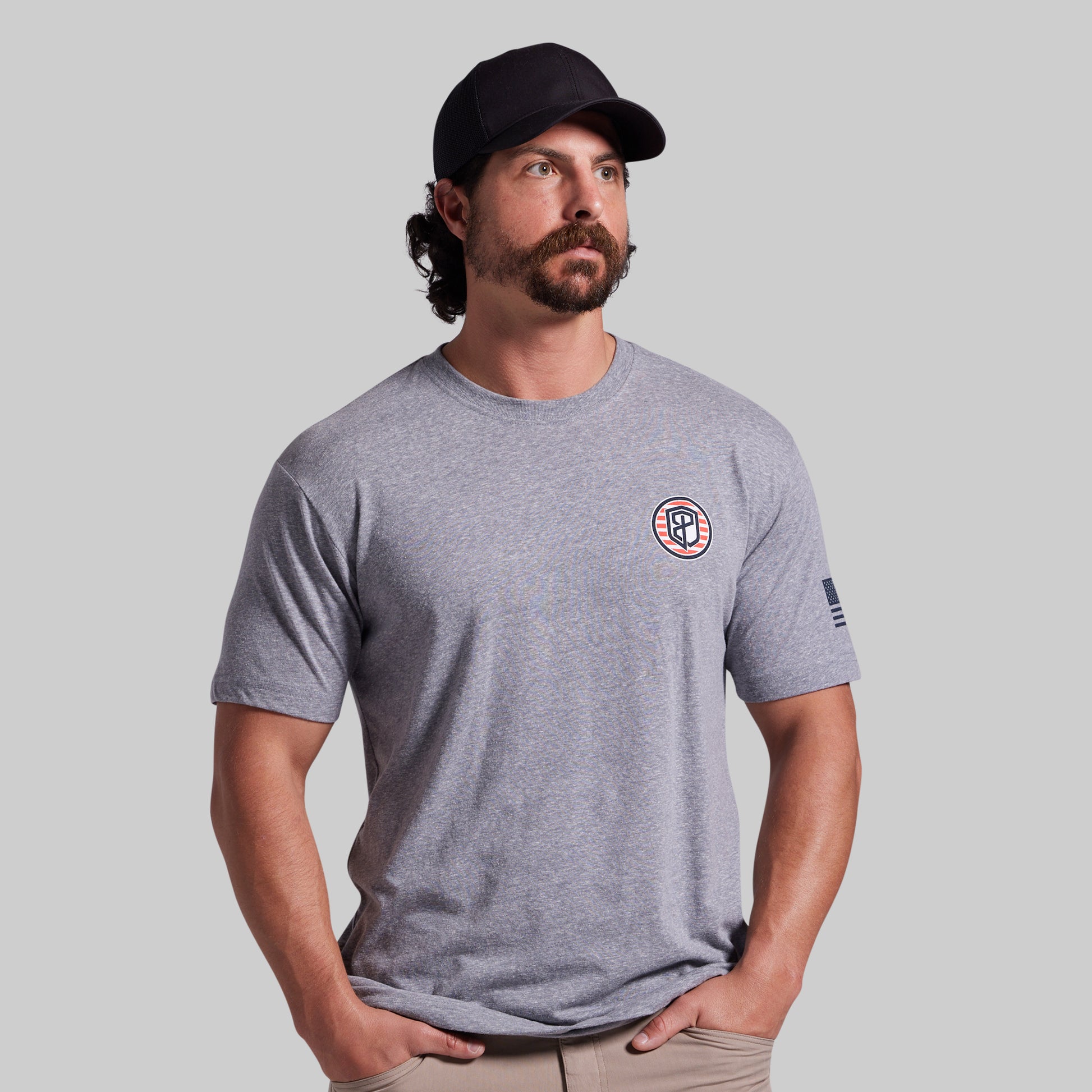 A man wearing a heather gray crewneck t-shirt with a circular logo on the left chest and a small flag print on the sleeve, paired with a black baseball cap and khaki pants, set against a solid gray background.