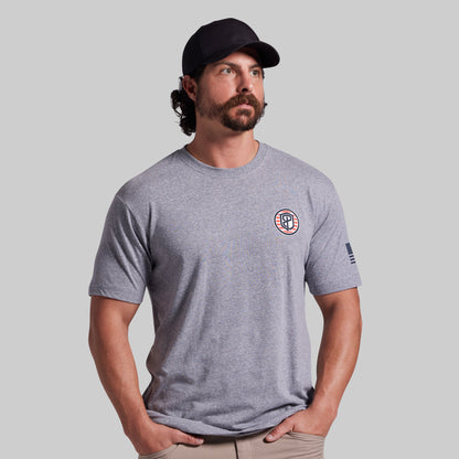 A man wearing a heather gray crewneck t-shirt with a circular logo on the left chest and a small flag print on the sleeve, paired with a black baseball cap and khaki pants, set against a solid gray background.