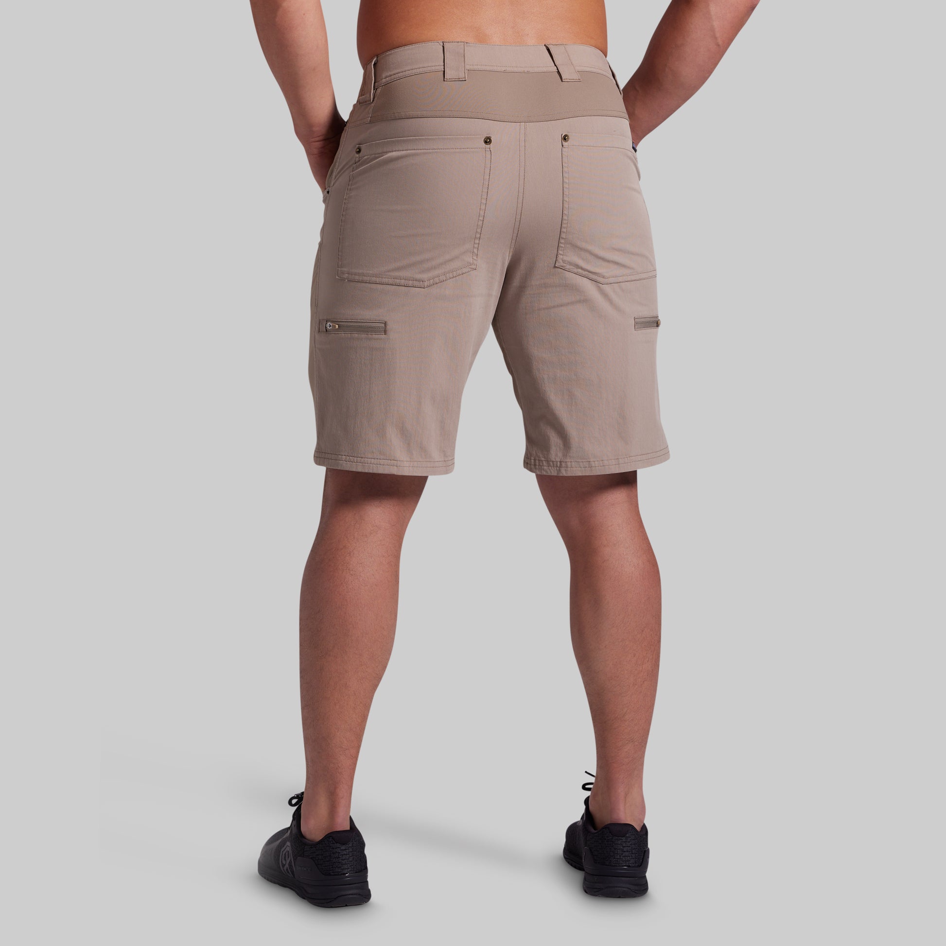 A man viewed from the back wearing beige cargo shorts and black sneakers against a solid gray studio background.