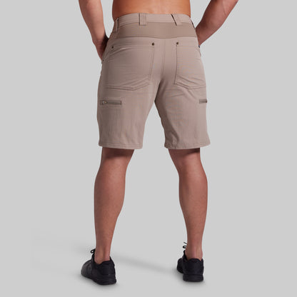 A man viewed from the back wearing beige cargo shorts and black sneakers against a solid gray studio background.