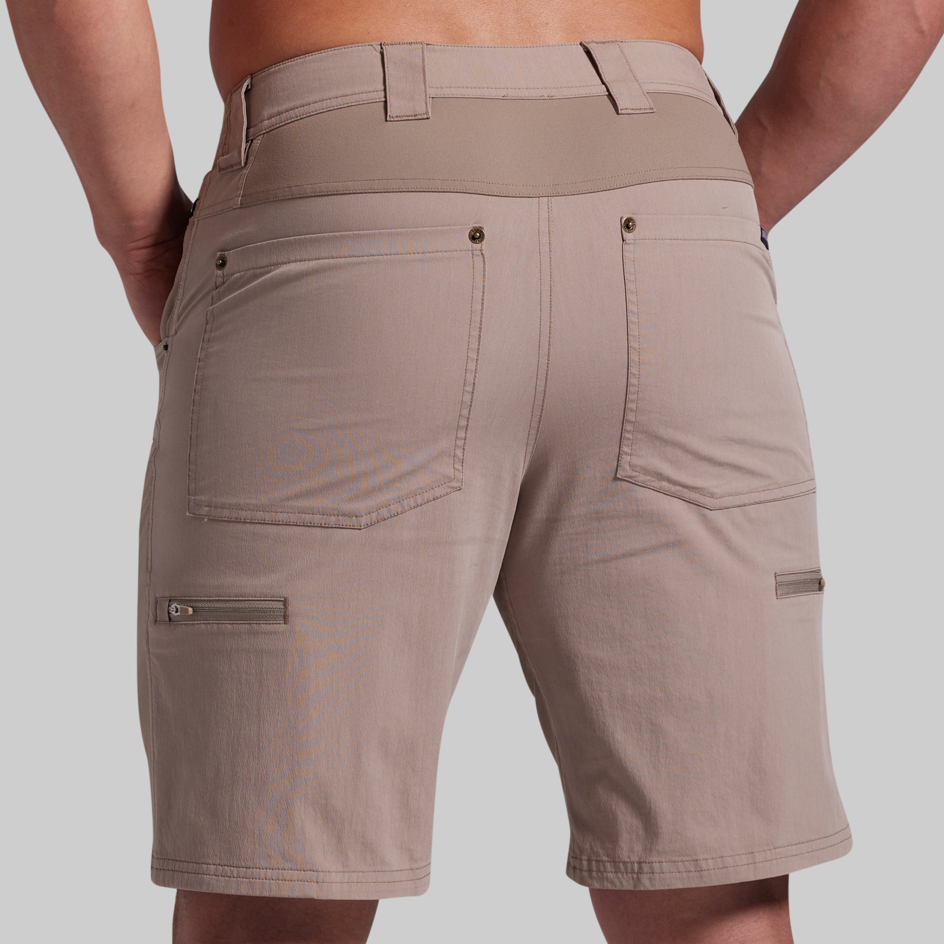 Rear view of a person wearing beige cargo shorts with a wide waistband, two large back patch pockets, and zippered side cargo pockets, photographed against a light gray studio background.