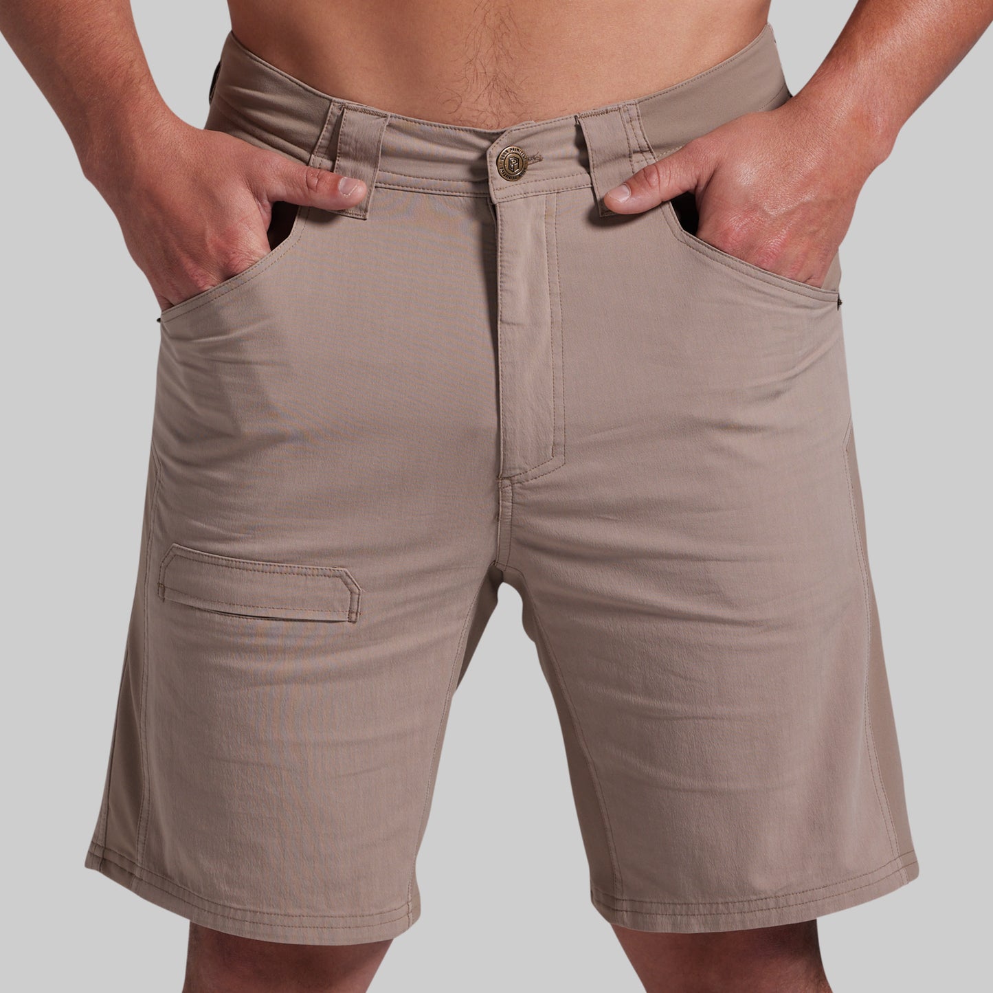 A close-up of a person wearing beige cargo-style shorts, with both hands in the front pockets and the waistband visible against a plain light gray background.