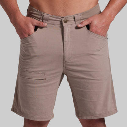 A close-up of a person wearing beige cargo-style shorts, with both hands in the front pockets and the waistband visible against a plain light gray background.