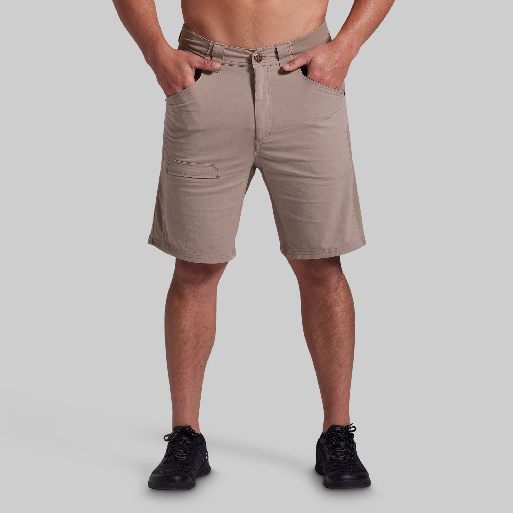 A man wearing khaki beige shorts with a relaxed fit, side pockets, and a button closure, paired with black sneakers, shown from the waist down on a solid gray background.