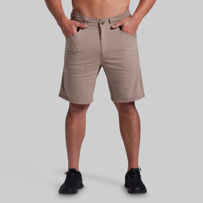 A man wearing khaki beige shorts with a relaxed fit, side pockets, and a button closure, paired with black sneakers, shown from the waist down on a solid gray background.