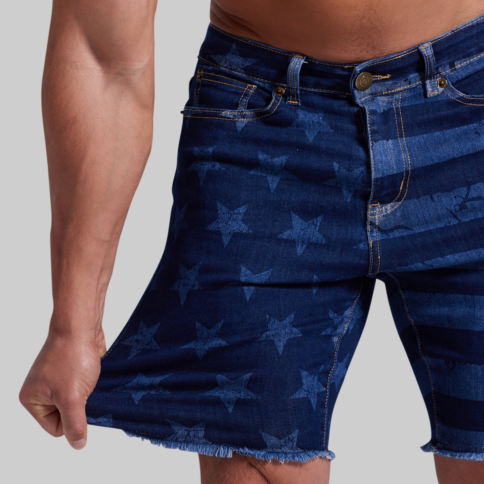 Close-up of a person wearing dark blue denim shorts with a tonal star pattern and frayed hems, shown against a light gray studio background.