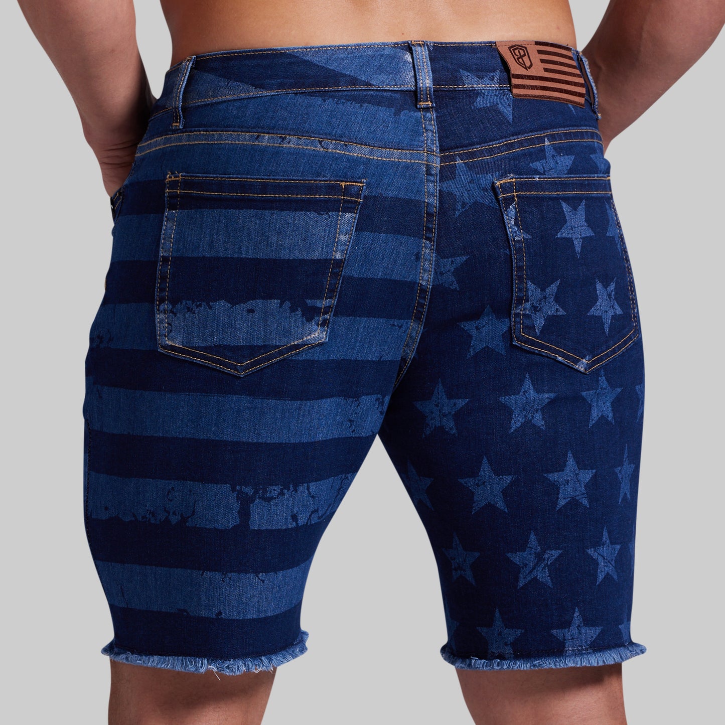 Rear view of a person wearing blue denim shorts with a distressed American flag pattern, featuring horizontal stripes and stars, frayed hems, and a brown leather patch on the back waistband.
