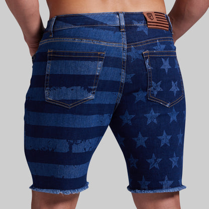 Rear view of a person wearing blue denim shorts with a distressed American flag pattern, featuring horizontal stripes and stars, frayed hems, and a brown leather patch on the back waistband.