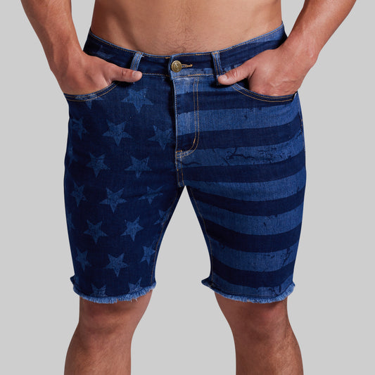 Men's denim shorts featuring a blue and navy American flag-inspired pattern with stars and horizontal stripes, shown on a model against a solid gray background.