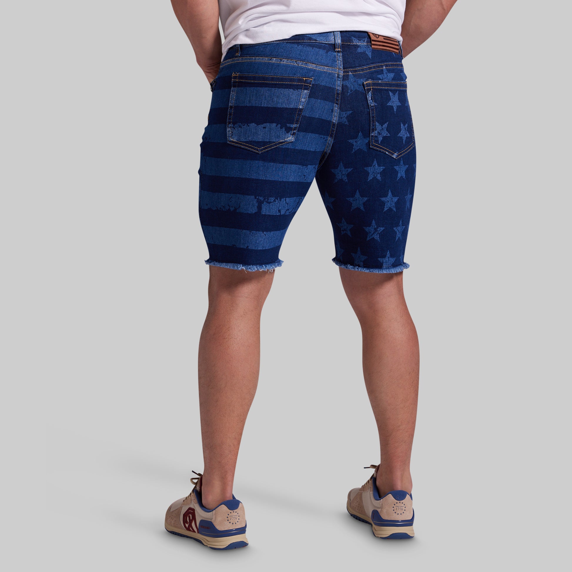 Rear view of a person wearing blue denim shorts with horizontal stripes on one leg and an allover star pattern on the other leg, paired with a white t-shirt and beige sneakers, against a solid gray background.