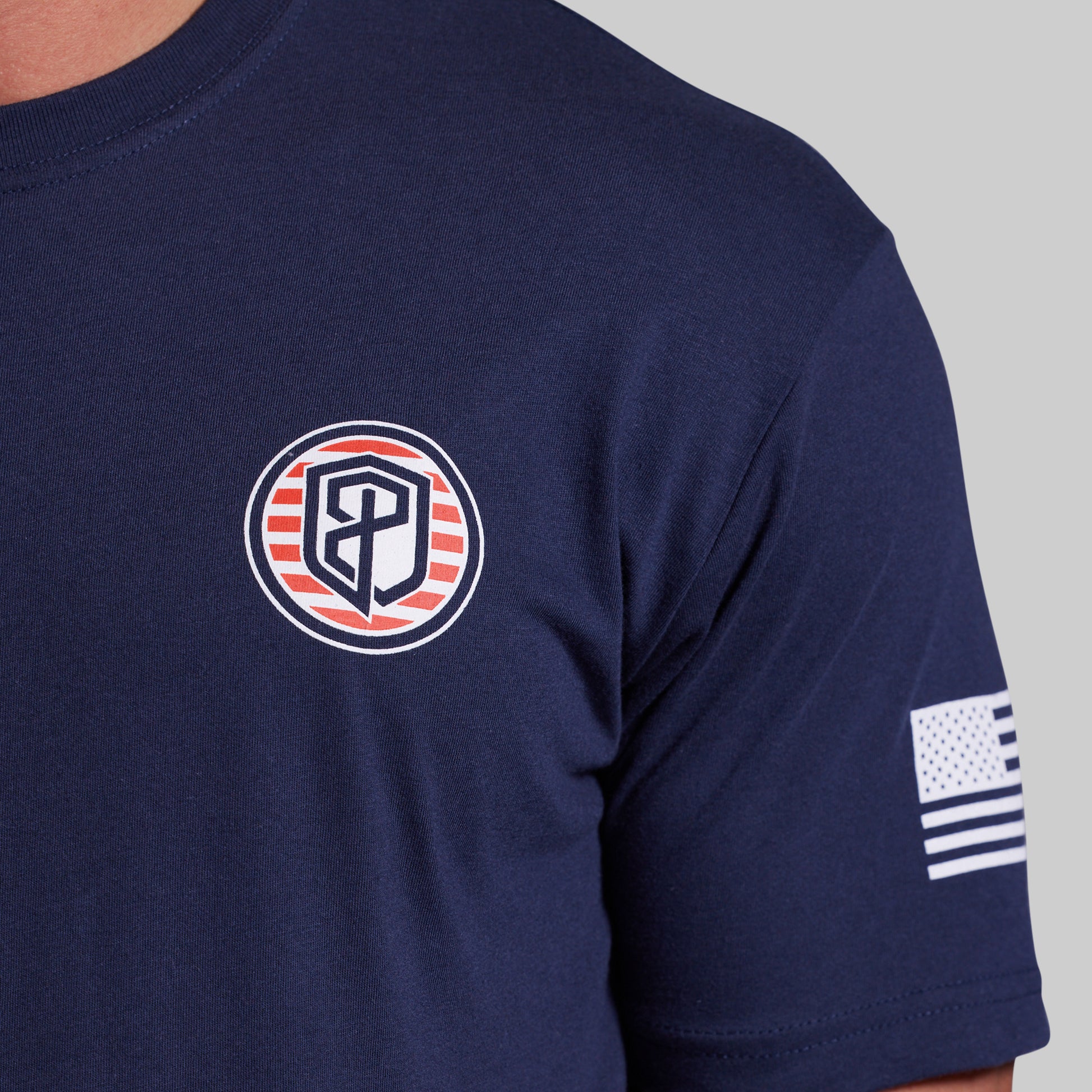 Close-up of a navy blue short-sleeve t-shirt worn by a model, featuring a circular chest logo with red and white stripes and a stylized monogram, and a small American flag patch on the sleeve.