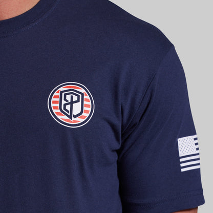 Close-up of a navy blue short-sleeve t-shirt worn by a model, featuring a circular chest logo with red and white stripes and a stylized monogram, and a small American flag patch on the sleeve.
