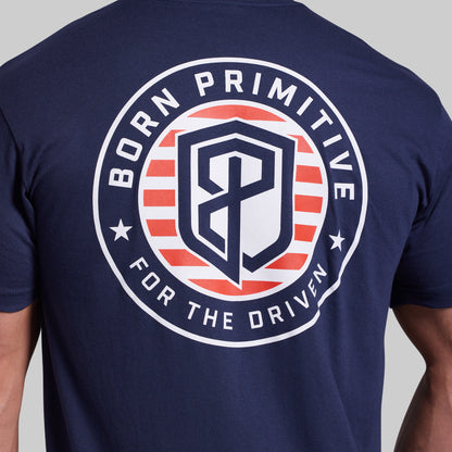 Close-up of the back of a navy blue t-shirt featuring a large circular graphic with red and white stripes, a stylized 'BP' shield, and the text 'BORN PRIMITIVE FOR THE DRIVEN'.