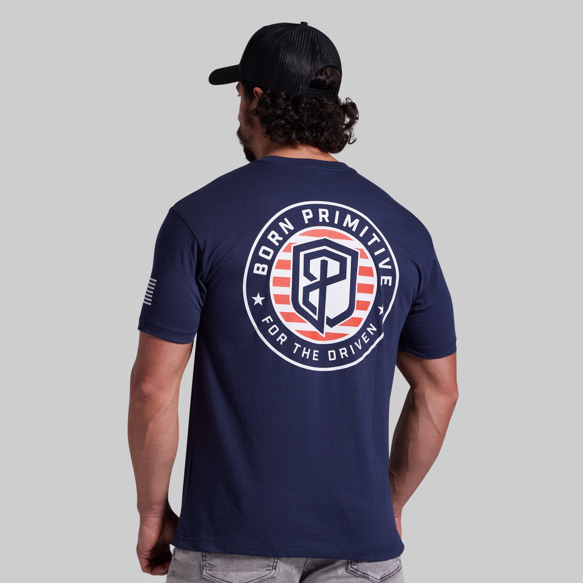 A man viewed from behind wearing a navy blue t-shirt with a large circular 'Born Primitive For The Driven' graphic on the back, paired with a black baseball cap and gray jeans, against a solid gray background.