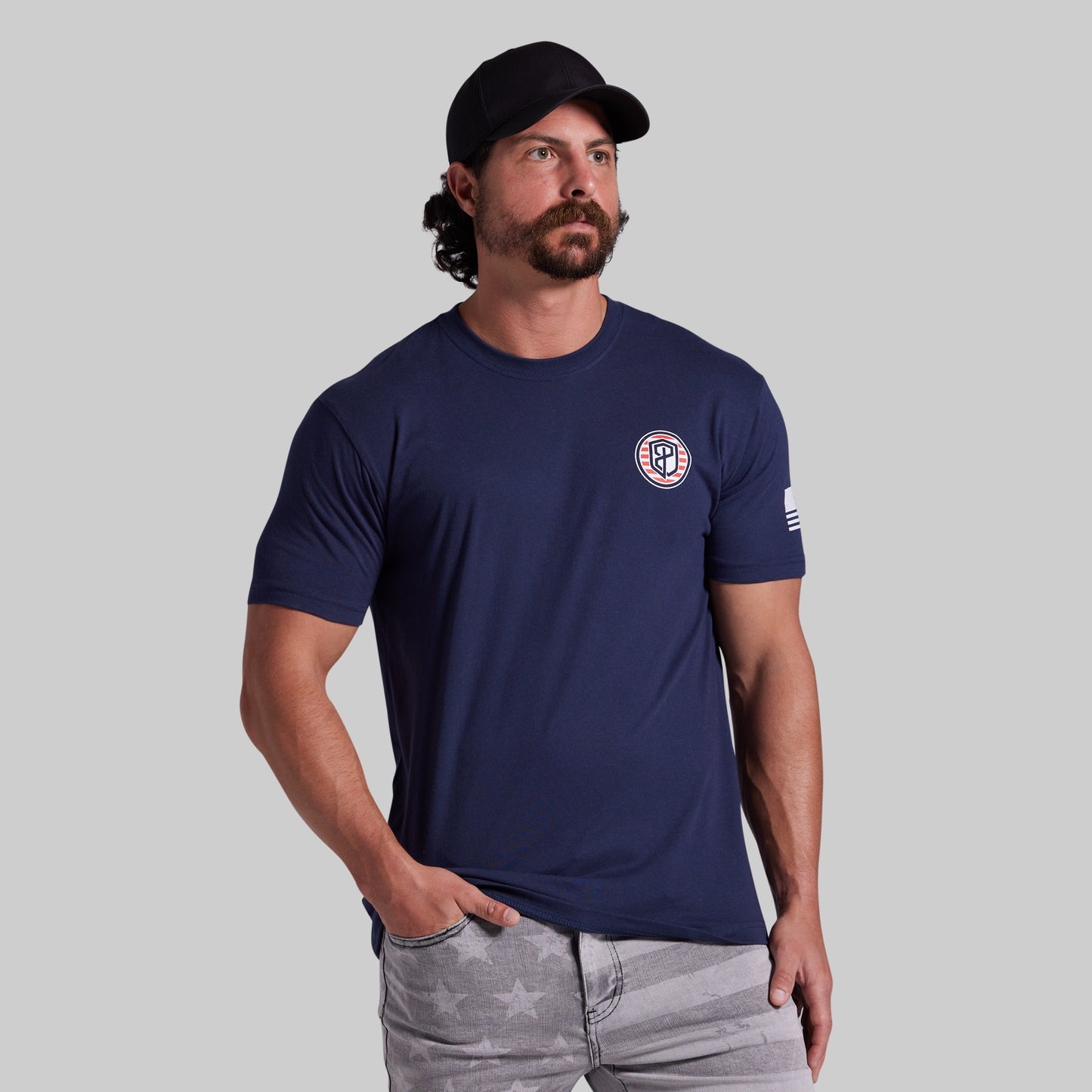 A man wearing a navy blue crewneck t-shirt with a circular logo on the left chest and a small flag graphic on the left sleeve, paired with gray jeans and a black baseball cap, set against a solid gray background.