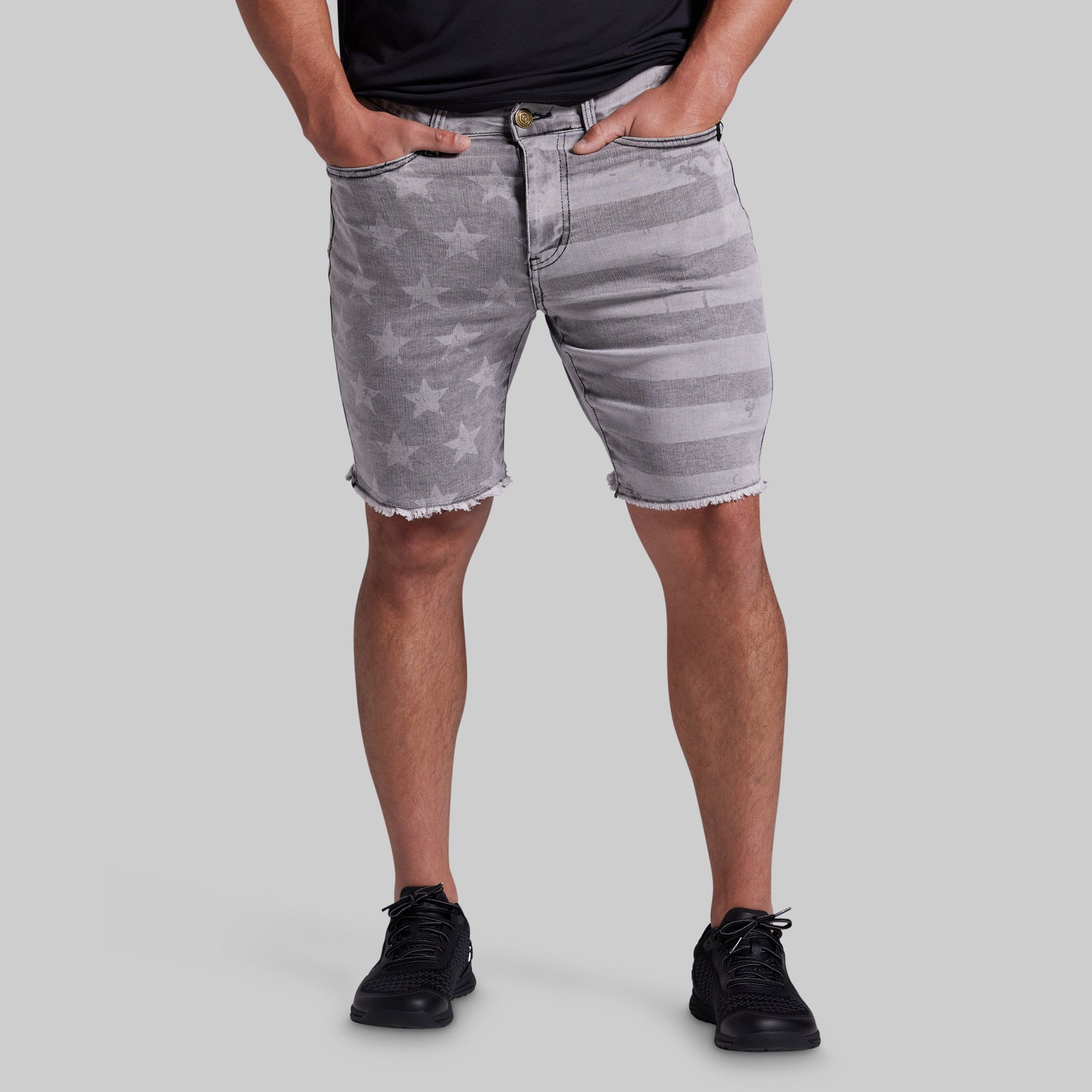 A person wearing gray denim shorts with a faded star pattern and horizontal stripe details, paired with a black shirt and black sneakers, against a solid gray background.