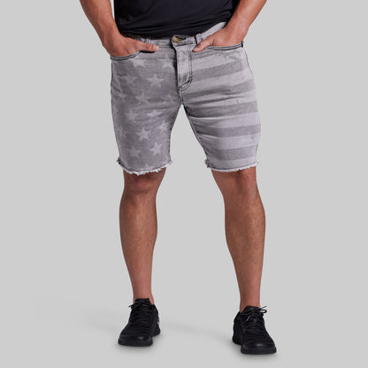 A person wearing gray denim shorts with a faded star pattern and horizontal stripe details, paired with a black shirt and black sneakers, against a solid gray background.