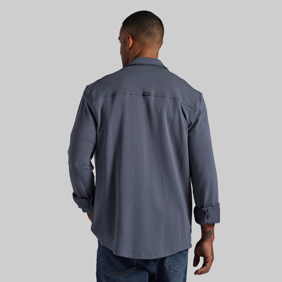 A man viewed from behind wearing a slate gray long-sleeve button-down shirt and blue jeans against a solid gray background.
