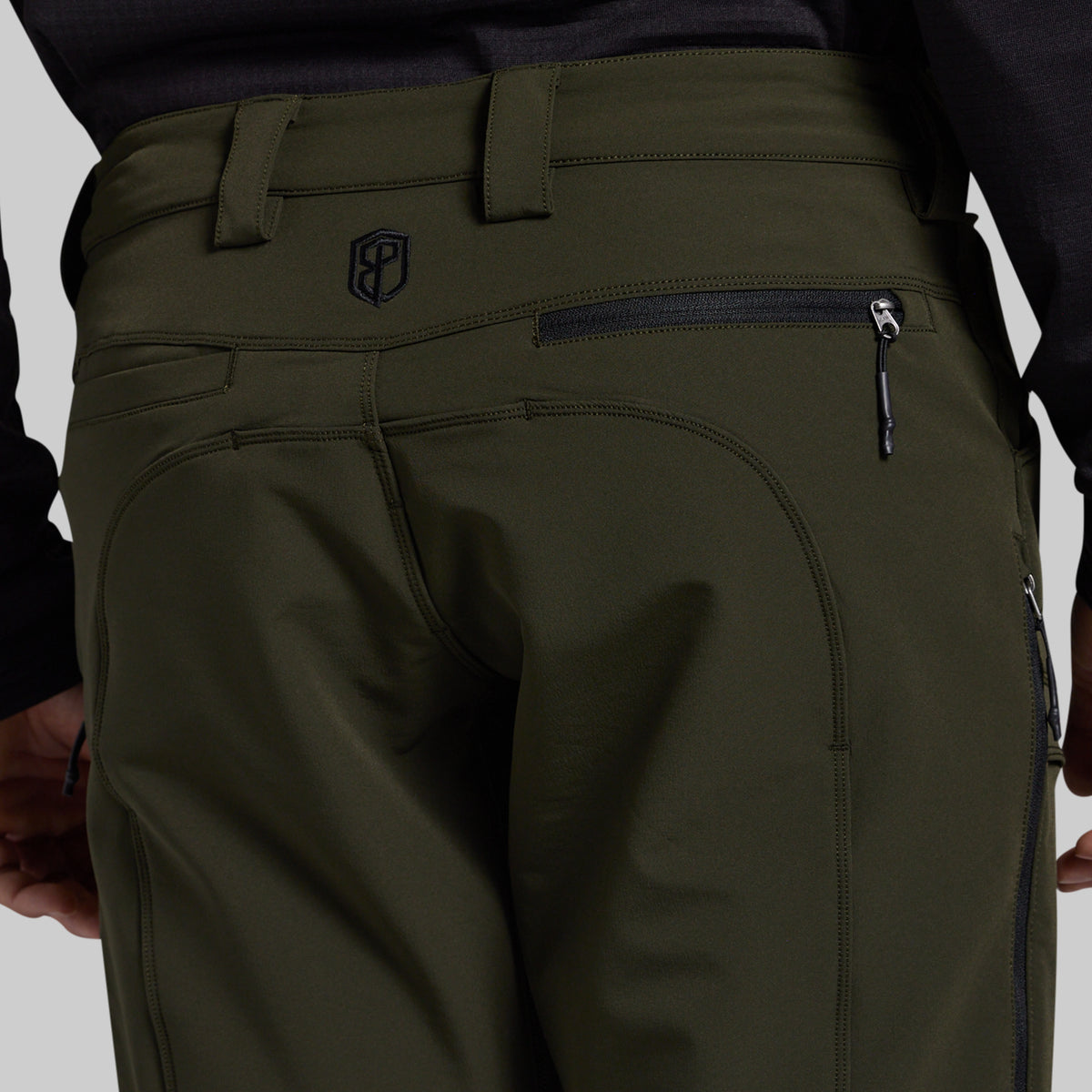 Close-up rear view of olive green outdoor pants worn by a model, showing the back waistband, belt loops, zippered pocket, and a small shield logo near the upper left side.