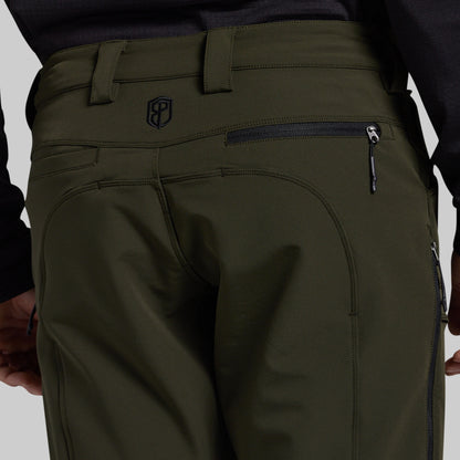 Close-up rear view of olive green outdoor pants worn by a model, showing the back waistband, belt loops, zippered pocket, and a small shield logo near the upper left side.