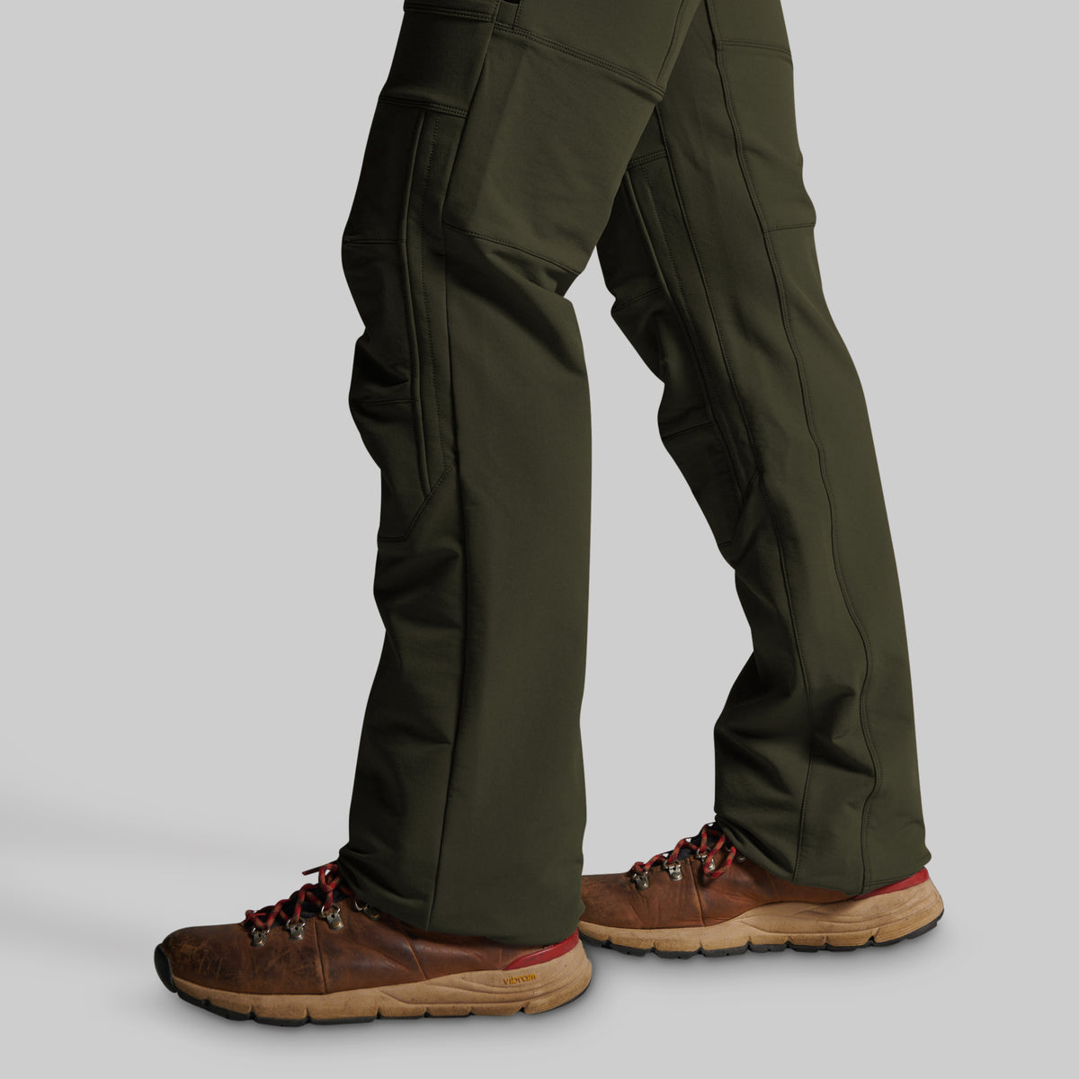 A close-up of a person's legs wearing dark olive green cargo pants and brown hiking boots against a solid gray background.