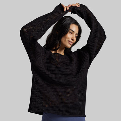 A woman modeling a black open-knit long-sleeve sweater against a solid gray background.