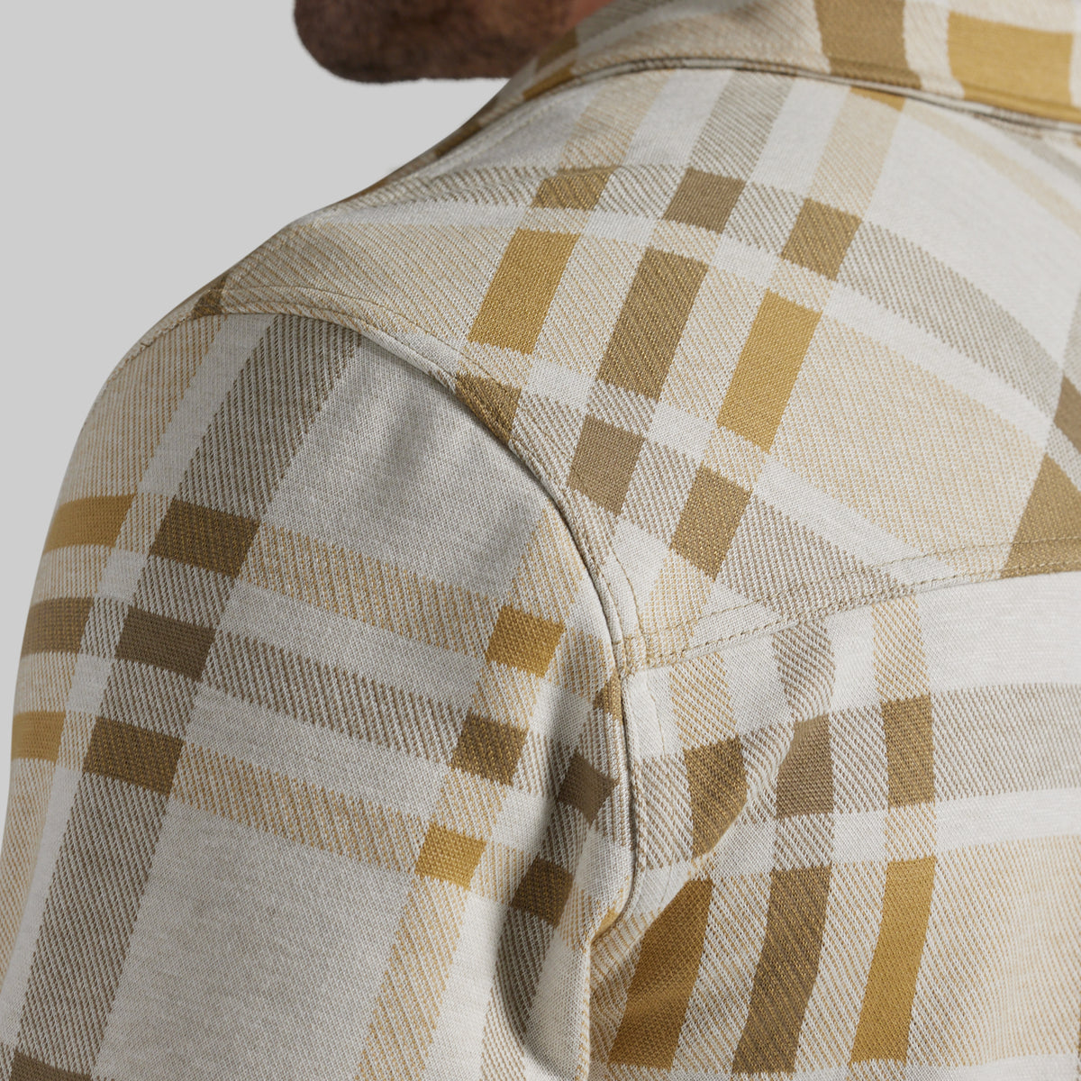 Close-up of a plaid flannel shirt on a model, showing the shoulder and upper chest. The pattern features a mix of beige, white, brown, and mustard yellow checks.