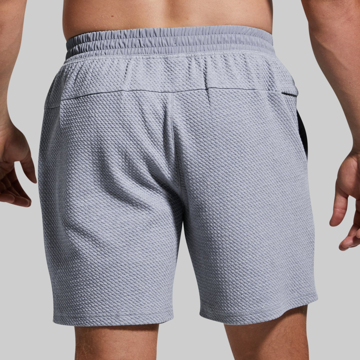 Rear view of a person wearing textured light gray athletic shorts with an elastic waistband and side pockets, shown against a plain light gray background.