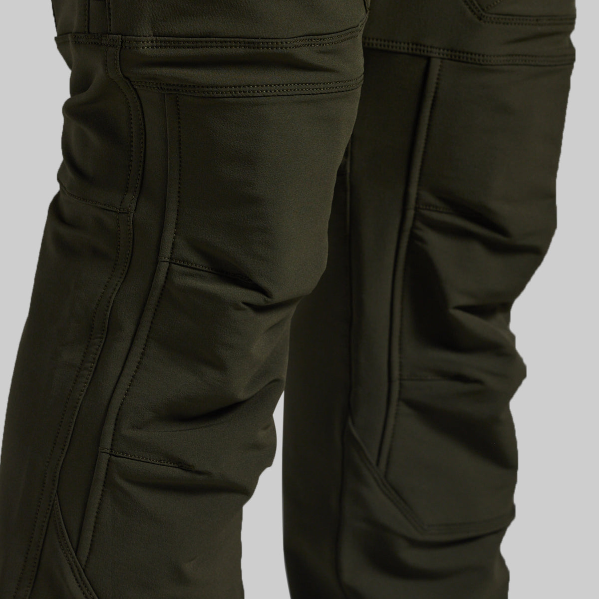 Close-up of dark olive green technical pants showing the leg construction, seam details, and pocket stitching against a neutral gray background.