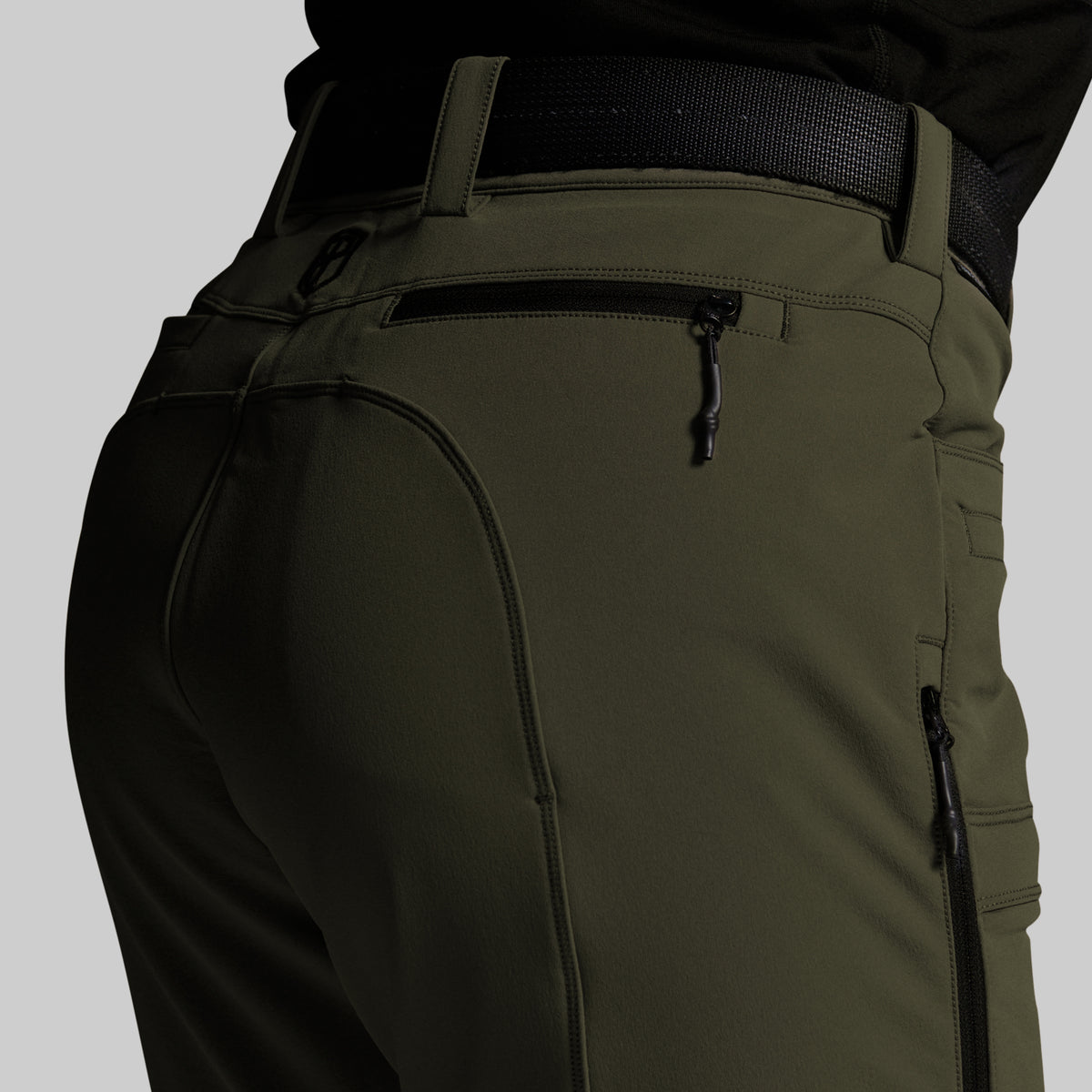 Close-up of olive green outdoor pants with a black belt, showing the back pocket, zipper details, and side cargo pockets on a neutral gray background.