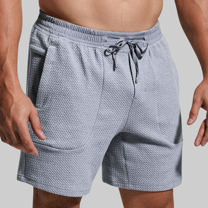 A close-up of a person wearing textured light gray drawstring shorts against a plain light gray background.