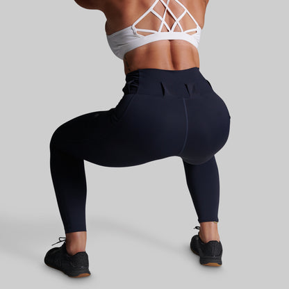 A woman wearing high-waisted navy blue athletic leggings and a white strappy sports bra, shown from the back while performing a deep squat against a solid gray background.
