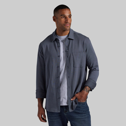 A man wearing a slate blue-gray button-up overshirt over a light gray t-shirt and blue jeans, standing against a solid gray background.