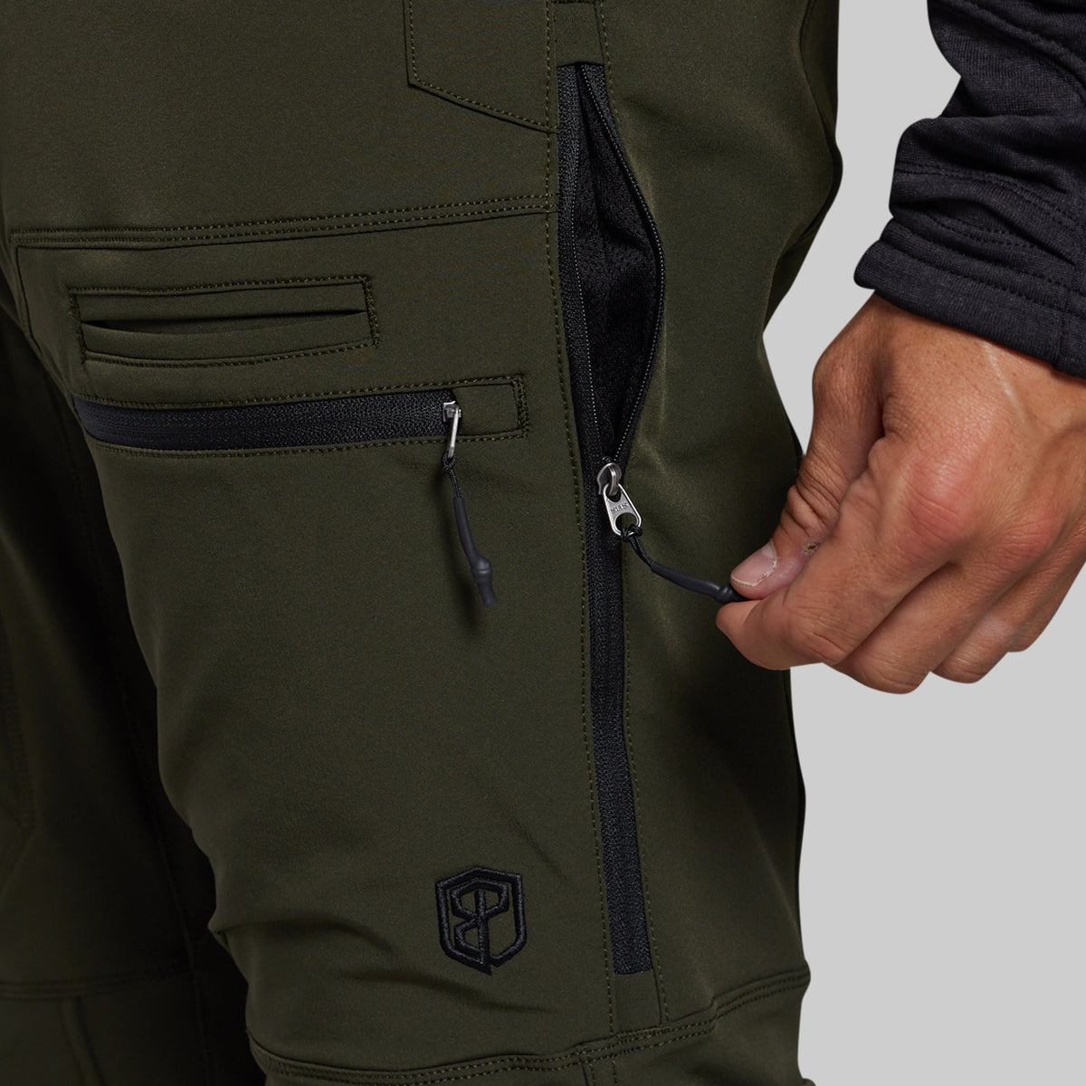 Close-up of olive green technical pants with a hand opening a zippered pocket, showing zippers and a small embroidered logo near the lower leg.