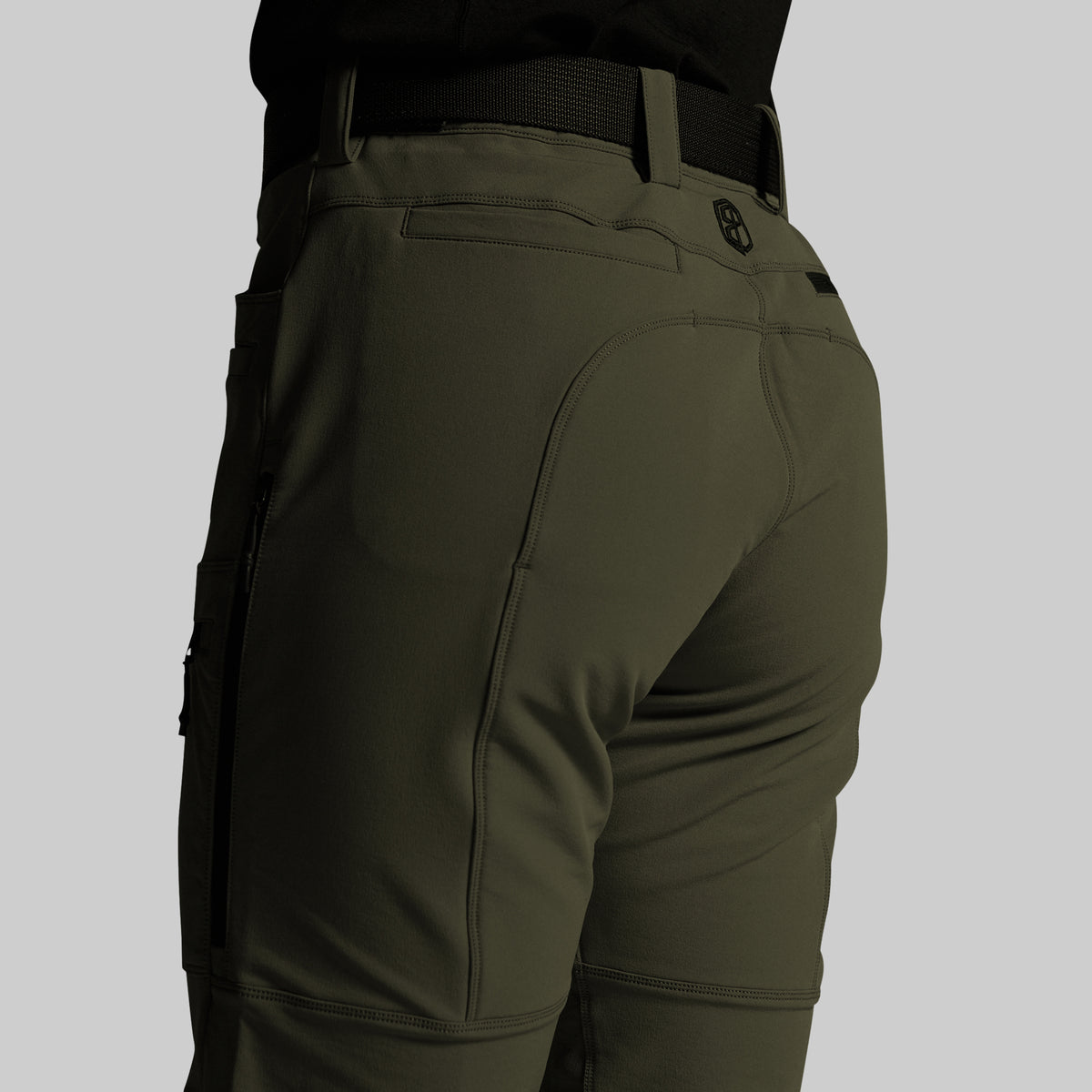 Close-up rear view of olive green outdoor pants worn by a model, showing the back pockets, belt loops, and a small circular logo near the upper right hip, against a light gray background.