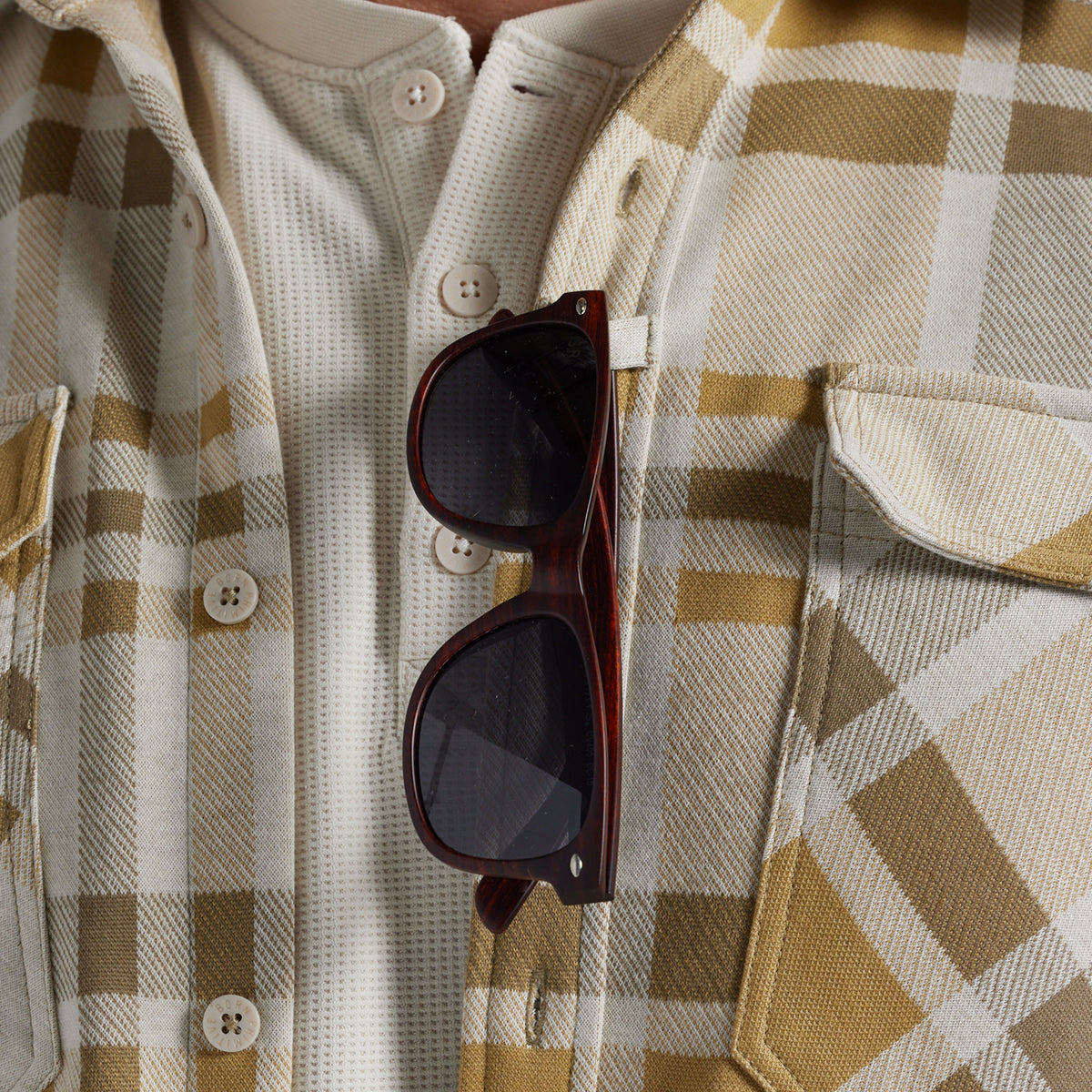A close-up of a person wearing a cream and tan plaid flannel shirt, with dark brown sunglasses hanging from the chest pocket.