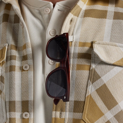 A close-up of a person wearing a cream and tan plaid flannel shirt, with dark brown sunglasses hanging from the chest pocket.