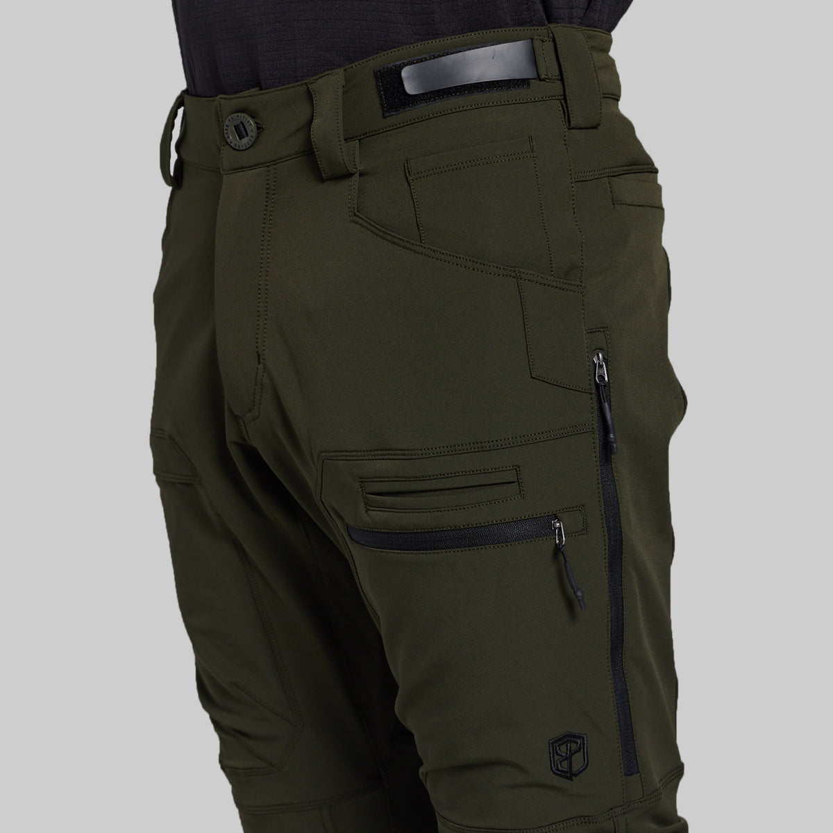 Close-up of olive green technical pants with multiple zippered cargo pockets, belt loops, and a dark belt, shown on a model against a light gray background.
