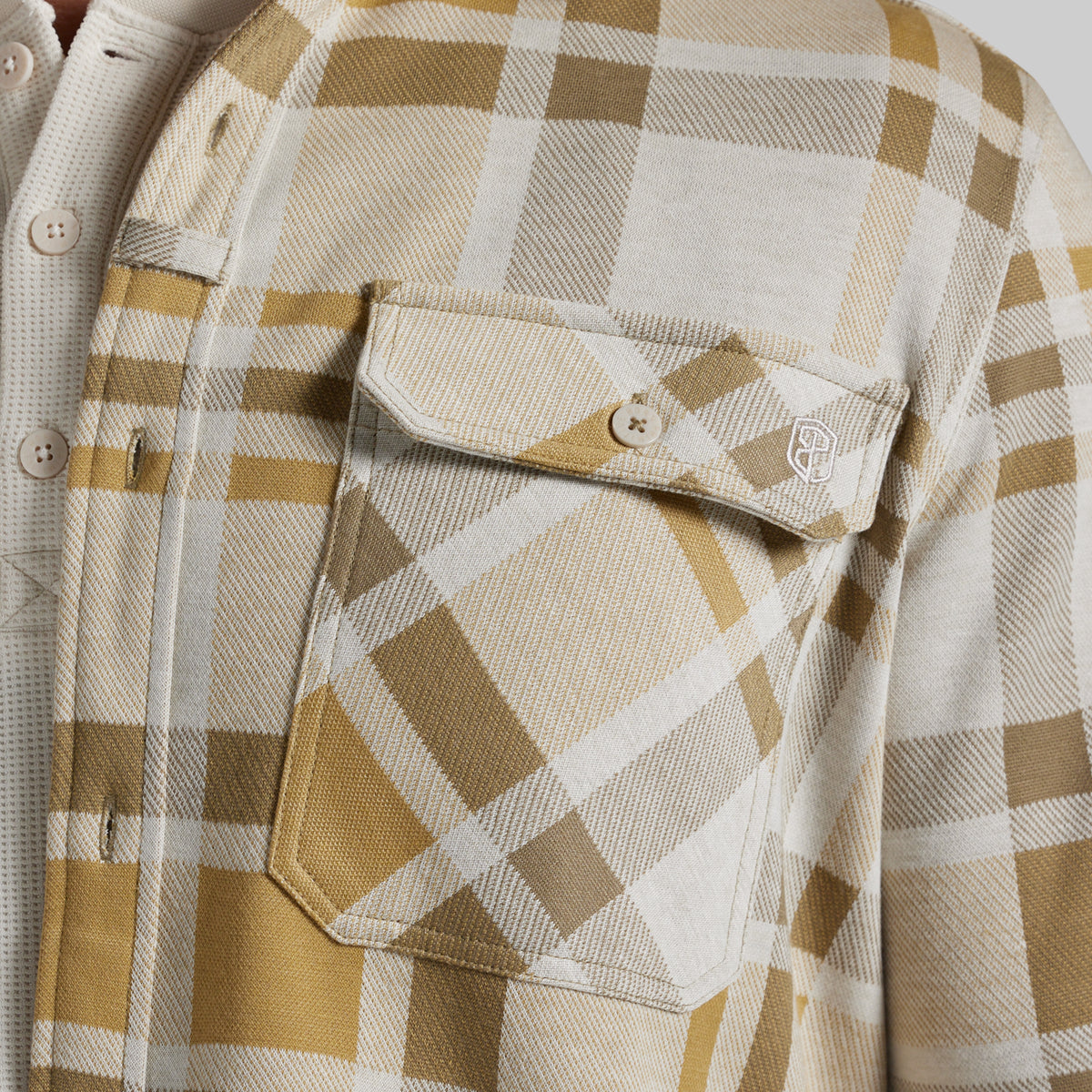 Close-up of a plaid overshirt with a chest pocket, featuring a pattern of tan, beige, and white. A small circular logo is visible on the pocket flap.