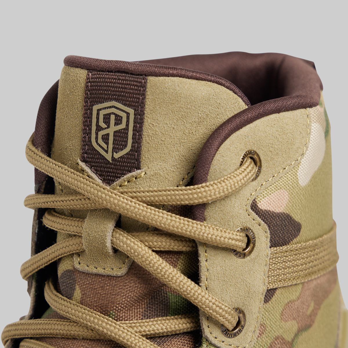 Close-up of the ankle collar of a tactical combat boot in a tan and brown camouflage pattern, featuring a dark brown tongue patch with a gold logo and tan laces.