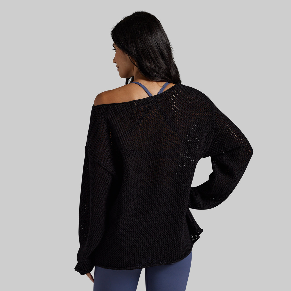 A woman shown from the back wearing a black off-the-shoulder mesh knit sweater over a blue strappy top and blue leggings, set against a solid gray background.