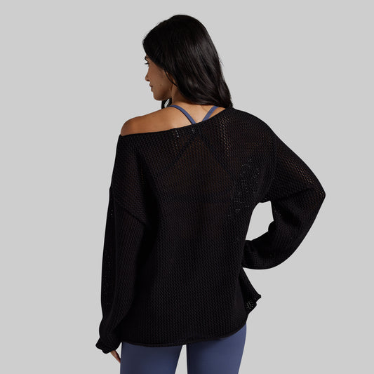 A woman shown from the back wearing a black off-the-shoulder mesh knit sweater over a blue strappy top and blue leggings, set against a solid gray background.