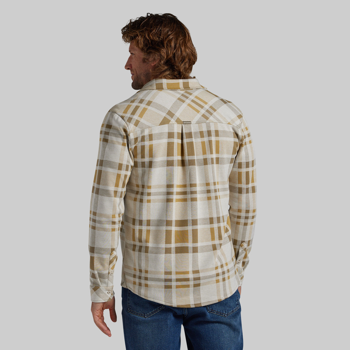 A man wearing a long-sleeve plaid flannel shirt in beige and brown tones, shown from the back against a solid gray background.